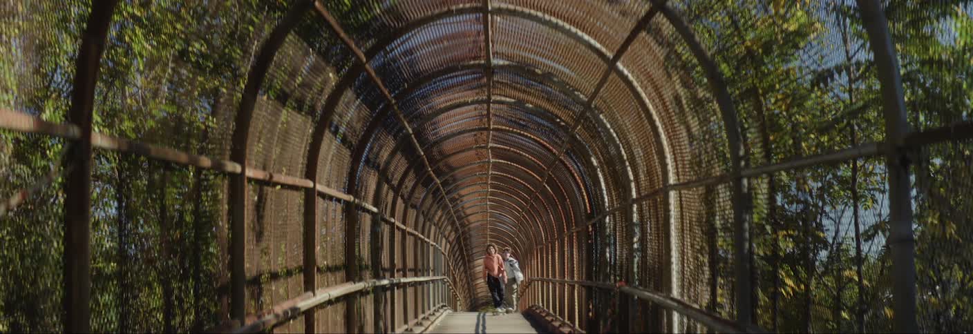 Two boys skateboarding through an arched hallway surrounded by trees