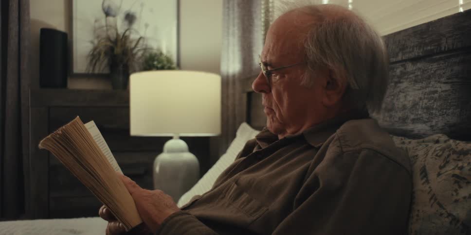An elderly man wearing glasses and a brown shirt sits on a bed in the bedroom reading a book and chuckling