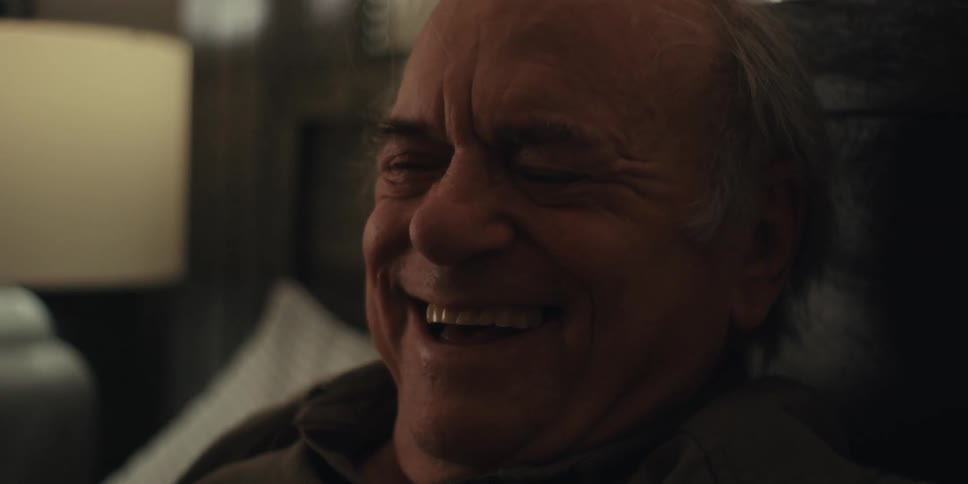 An older man reclining on a bed reading a book laughs and takes off his glasses in the bedroom