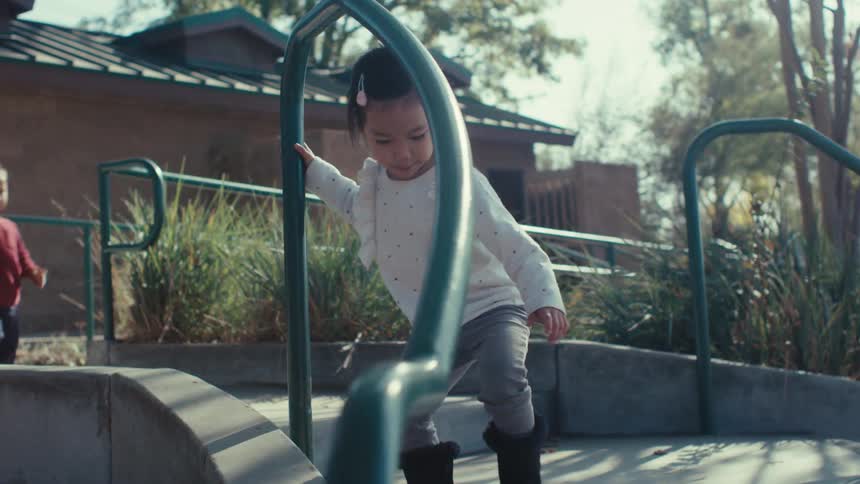 A child wearing a white sweater and gray pants walks down a curved concrete ramp with green metal railings in an outdoor park area