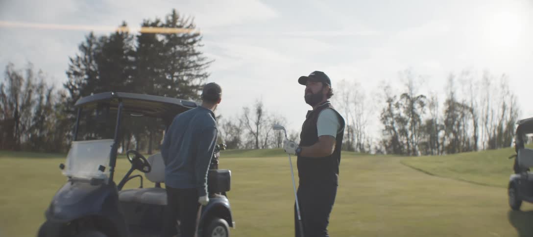 Two men wearing caps and casual golf attire exchange a fist bump beside a golf cart on a sunny day with trees in the background