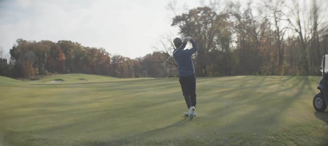 A golfer who has just hit the ball on a sunny day standing on a well manicured golf course with lush green grass surrounded by trees displaying autumn foliage under a bright clear sky