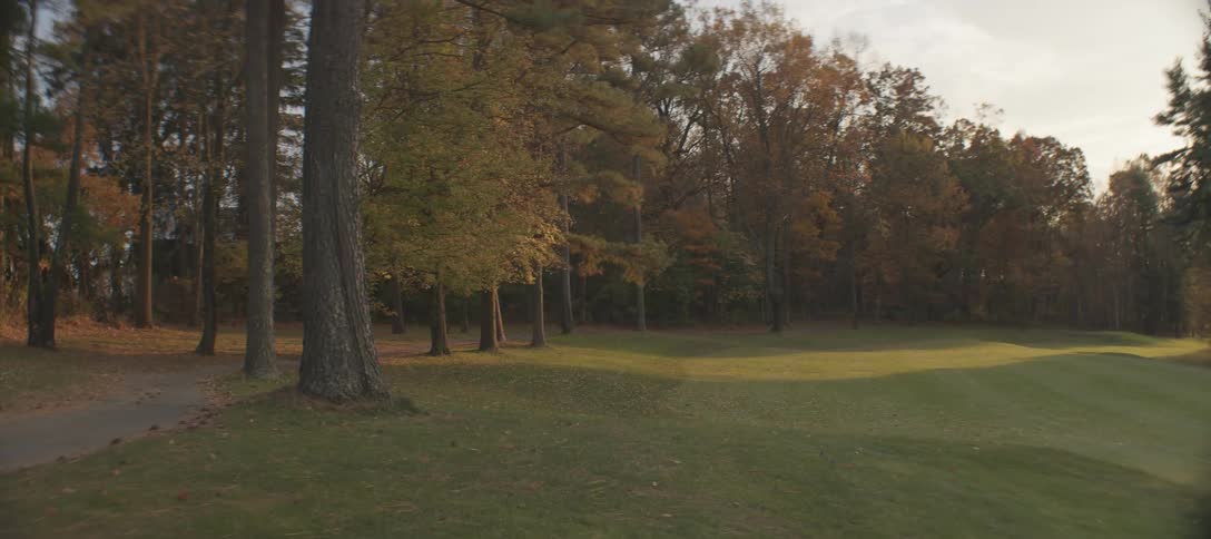 A golf cart is driving on a paved path next to a golf course surrounded by tall trees with autumn foliage