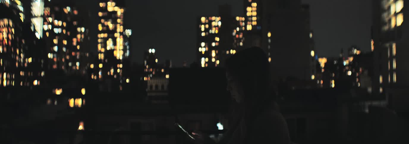 A woman using a smartphone is silhouetted against the illuminated tall buildings at night