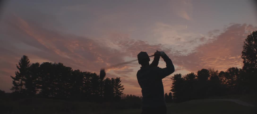 A golfer striking the ball with a club is silhouetted against the setting sun on the golf course