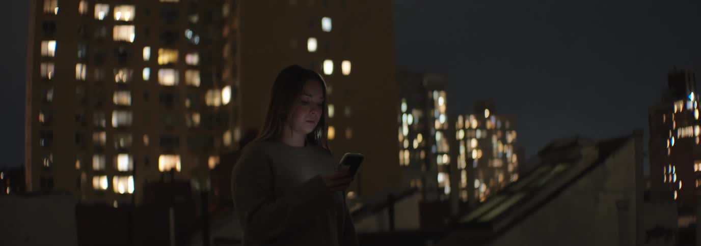 A woman is walking on a rooftop at night holding a smartphone with illuminated highrise buildings in the background