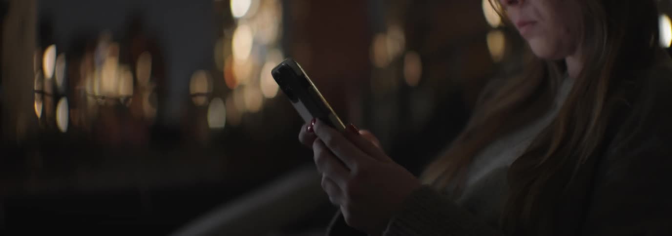 A woman with brown hair uses a smartphone with illuminated urban buildings in the background