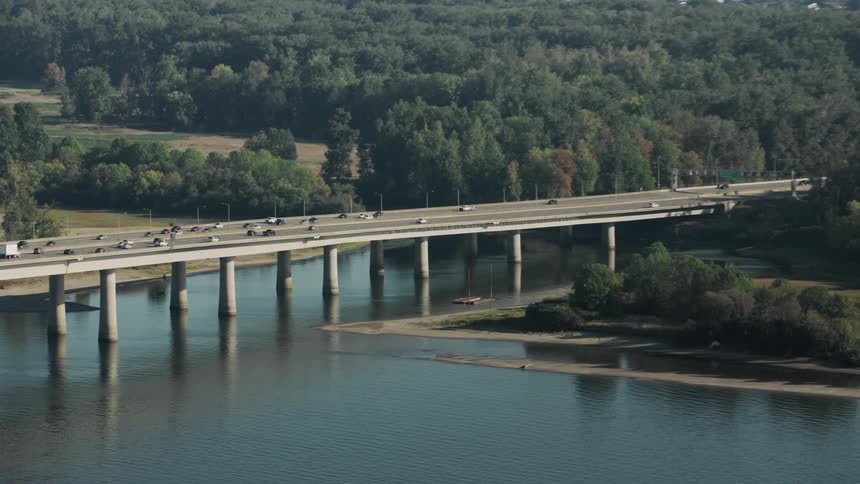 An aerial view of a long bridge spanning a wide river with traffic moving in both directions and lush greenery surrounding the waterway