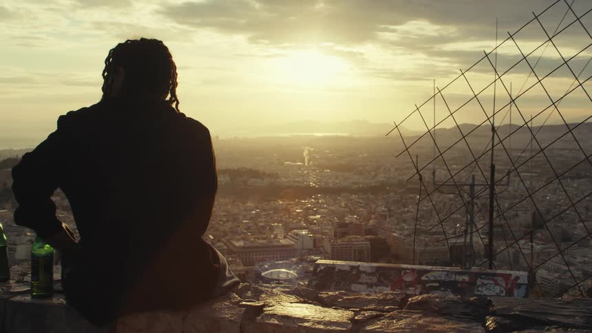 A person with braided hair sits on a ledge overlooking a sprawling cityscape at sunset