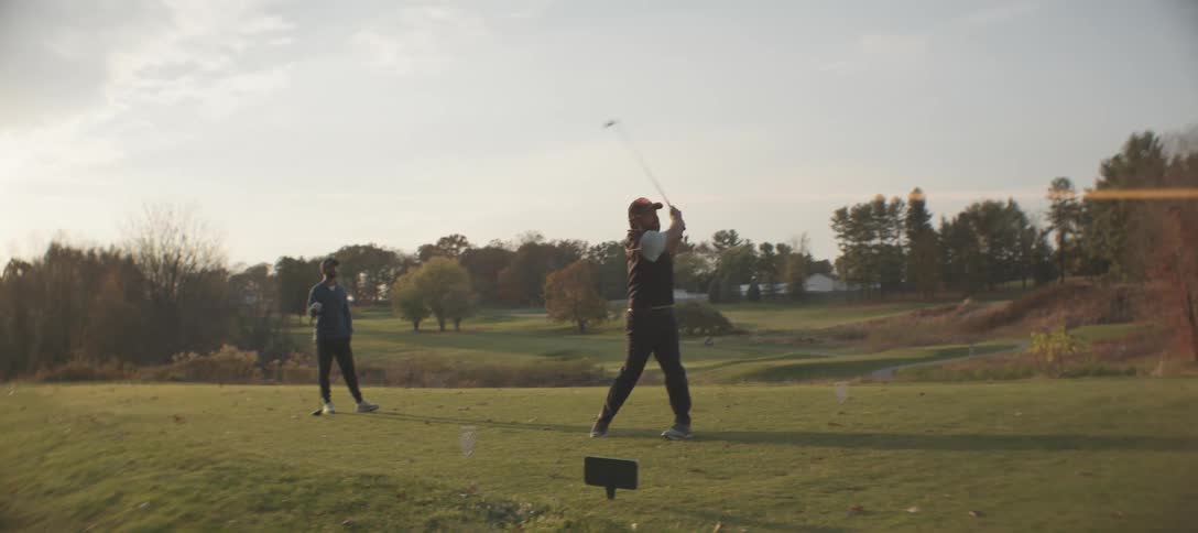 A golfer is striking the ball on a lush golf course while another person stands nearby watching surrounded by open greenery and trees under a calm cloudy sky