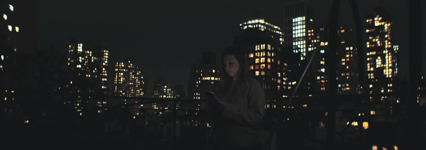 Woman stands outdoors and uses a device while lights shine from the downtown skyscrapers in the background at night