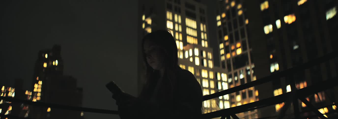 Woman using a smartphone is silhouetted against the illuminated urban buildings in the city at night