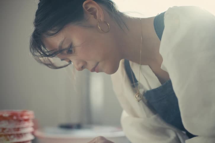 A potter wearing a blue apron and a white shirt carefully shapes a wet grey clay pot on a rotating pottery wheel