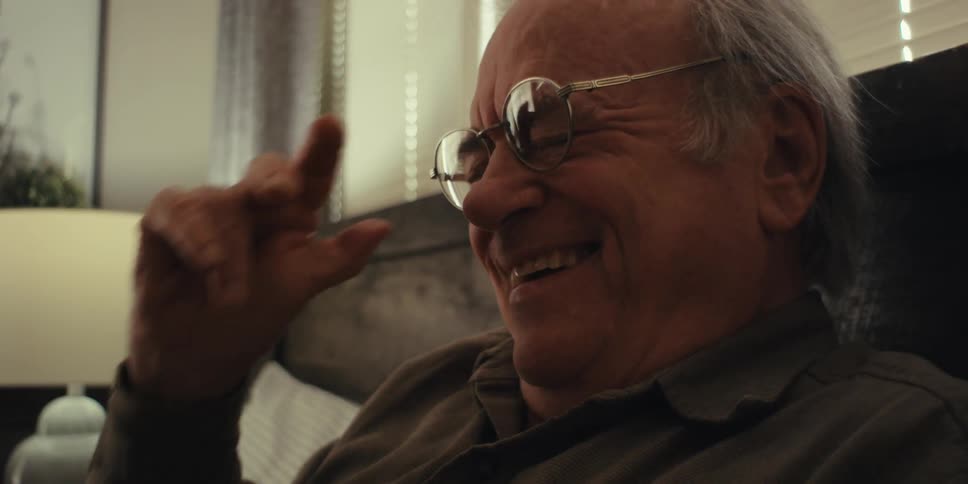 An elderly man in a blue shirt reads a book takes off his glasses and laughs near the table lamp in the bedroom