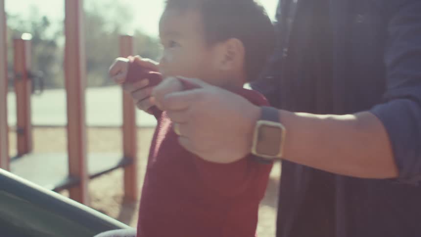 Father wearing a blue shirt and a wristwatch helps toddler son climb up a slide on the playground