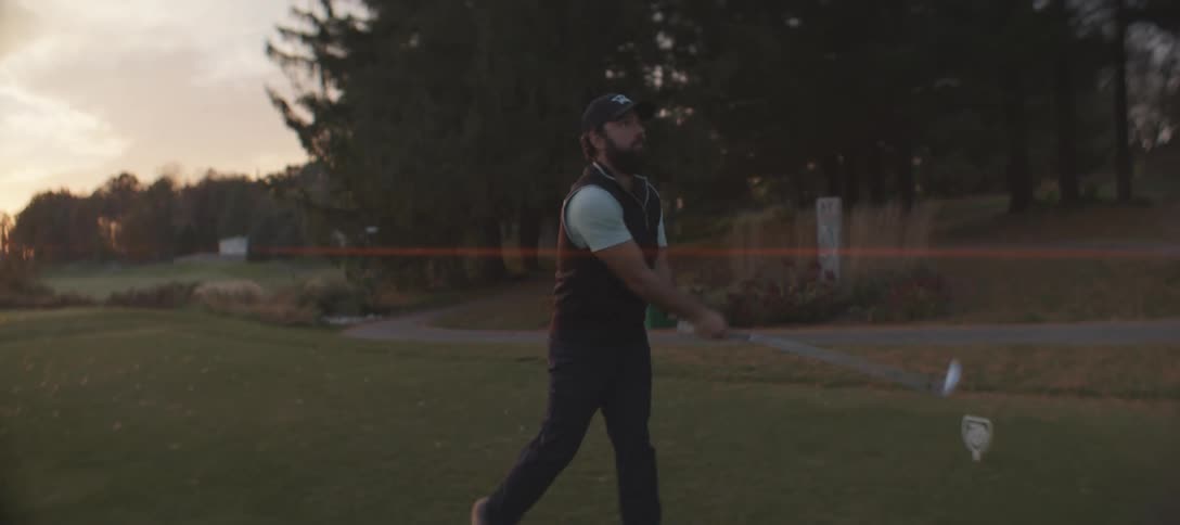 A golfer with a beard wearing a black vest a white shirt and pants strikes a golf ball with a golf club on the course