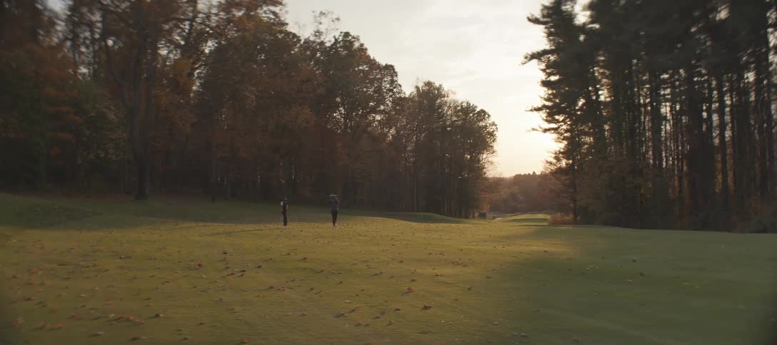 Two people stand on a golf course surrounded by autumnal trees under a partly cloudy sunset sky