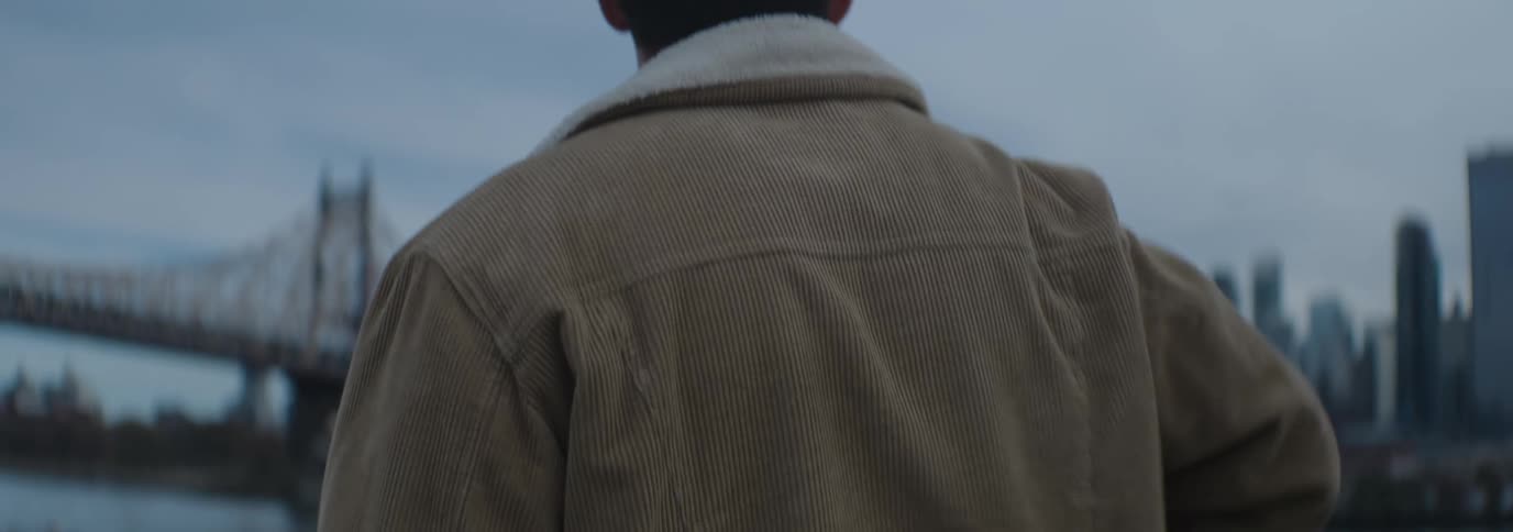 A man wearing a brown jacket with a white fleece collar walks along a riverside under a cloudy sky
