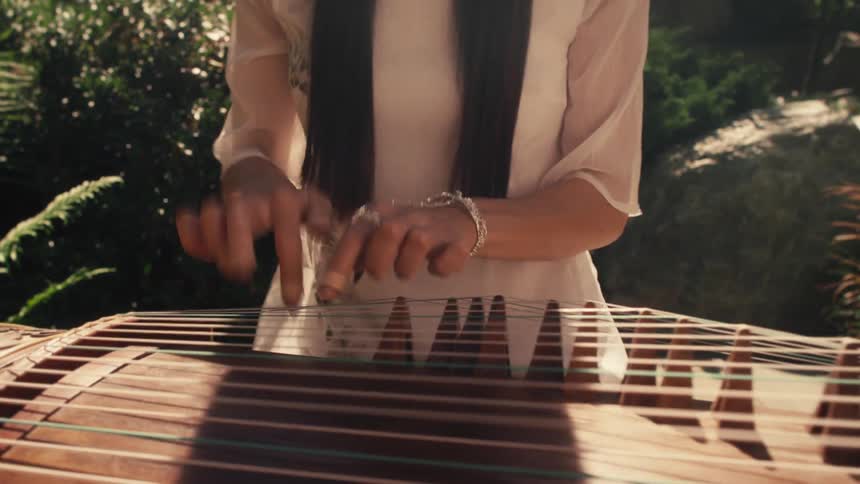 In a lush garden bathed in soft sunlight a woman dressed in flowing traditional attire gracefully plays a guqin her delicate fingers gliding over the strings as vibrant greenery and elegant stonework frame the tranquil scene