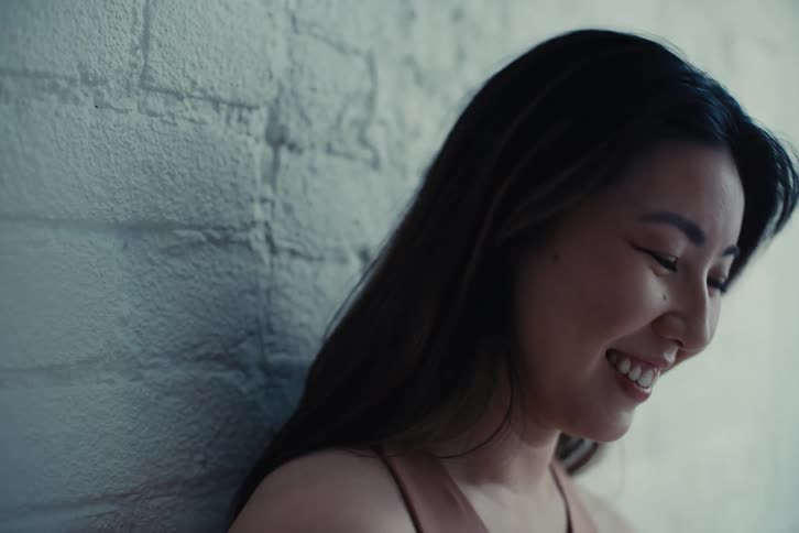 A woman with straight hair and a crop leans against a white brick wall laughing