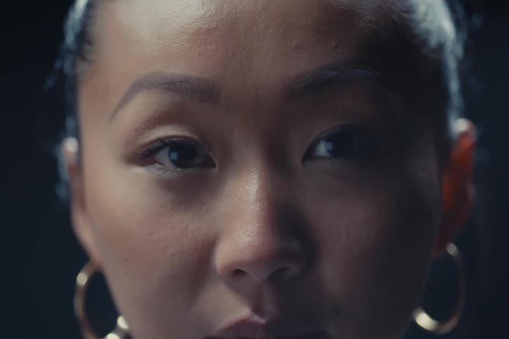 A womans face in low light emphasizing her eyes eyebrows natural skin texture and gold hoop earrings against a dark blurred background