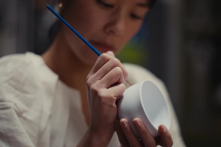 A person holds a white bowl in one hand and a paintbrush in the other painting it