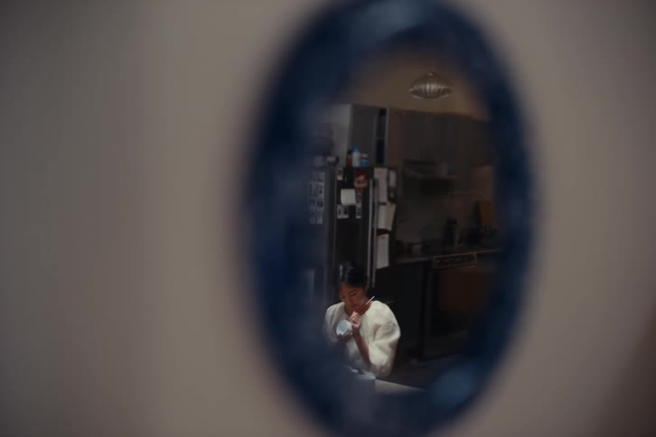 A woman shown through a small circular window is painting mugs in the kitchen