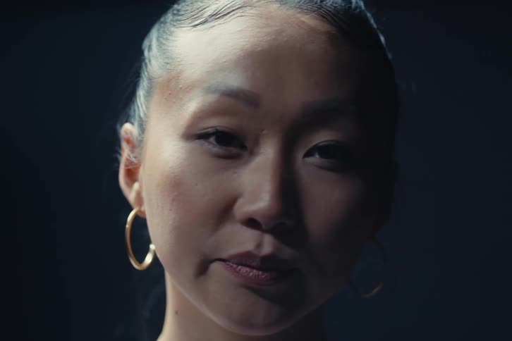 A smiling womans face in low light emphasizing her eyes eyebrows natural skin texture and gold hoop earrings against a dark blurred background