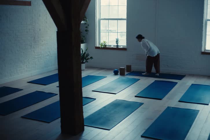 Woman in a room walking to a blue mat and stretching her body