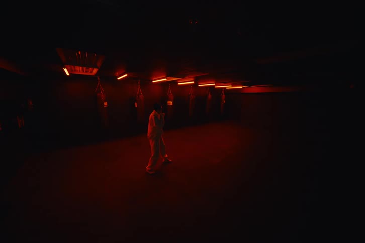 A person in a dark room illuminated by red light practices martial arts in front of hanging punching bags