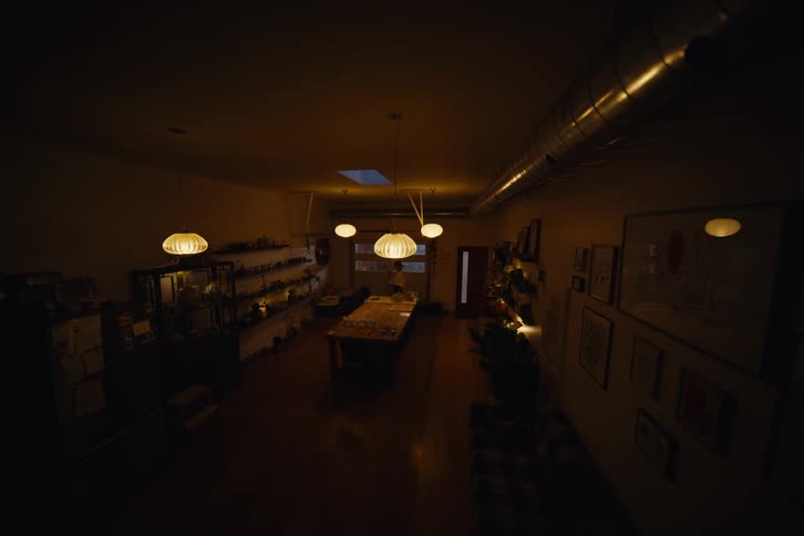 A person enters a dimly lit workshop where a long rectangular table is covered in tools and materials with a row of hanging light fixtures casting a warm glow overhead