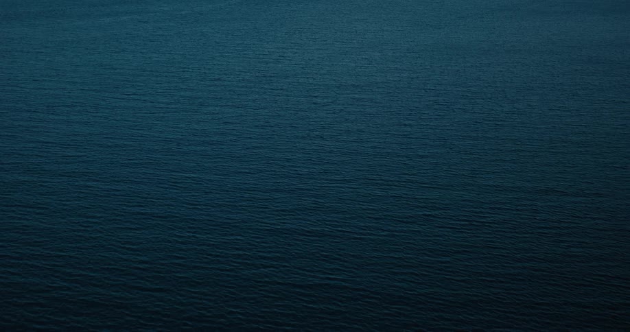 A dark rocky coastline with waves crashing against the rocks overlooking a vast blue ocean