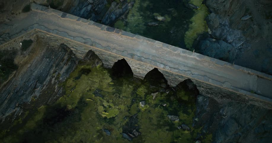An aerial view of a weathered stone dam with a narrow road running along its top spanning a river with dark murky green water