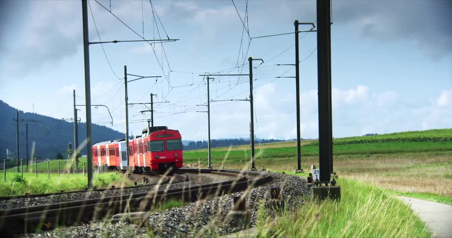 A red train is traveling on a curved railway track through a grassy rural landscape with overhead electric lines and utility poles