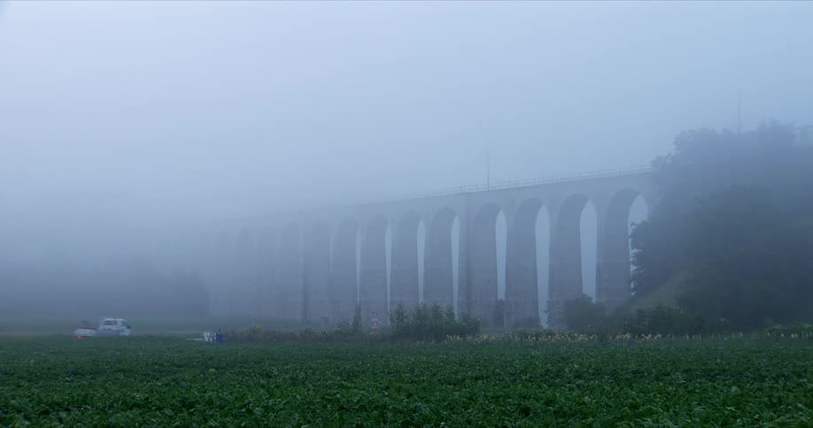 A foggy scene shows a long stone viaduct with multiple arches spanning across a field of crops