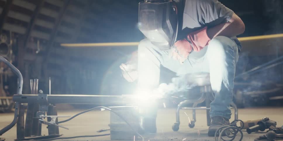 A man sitting down wearing a welding helmet to weld a piece of iron that is giving off several bright sparks