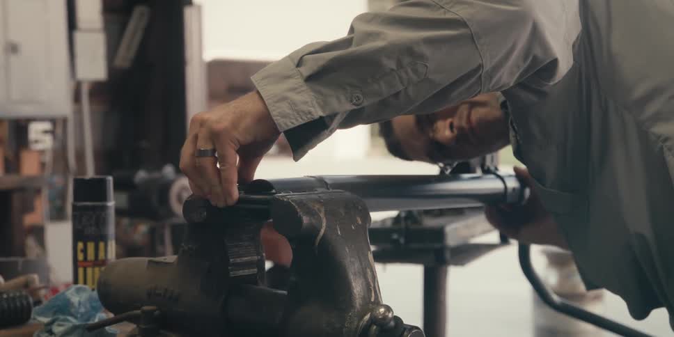 A man in longsleeved clothing is clamping a piece of iron in a vise table