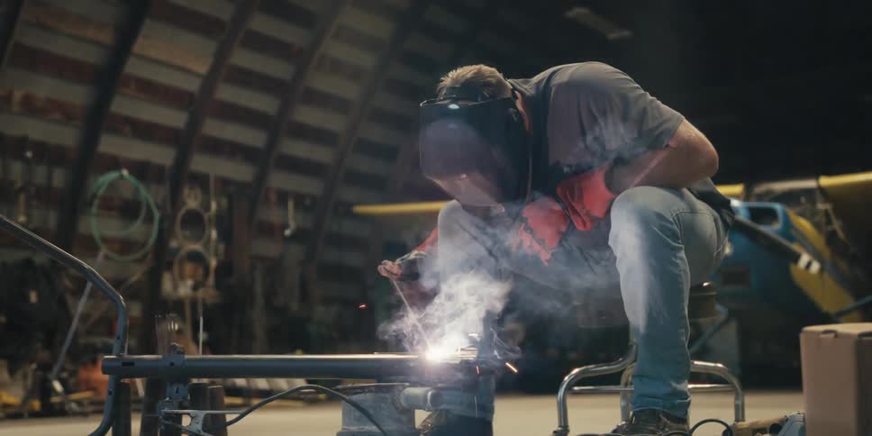 A man in a welding helmet is sitting and performing a bright weld and releasing several sparks in front of him