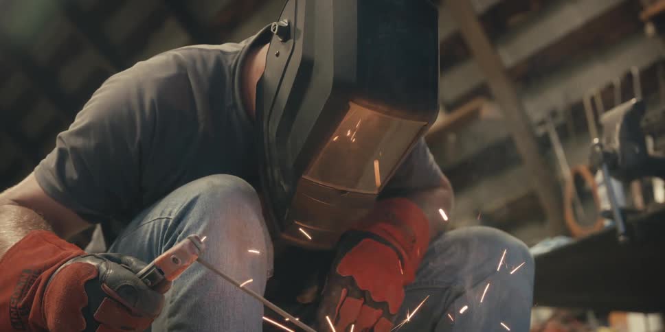 A person crouching down and wearing a welding helmet to weld a piece of metal and there are several sparks being thrown