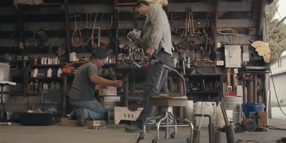 A man in a welding helmet is crouching on the ground while a man is walking with an iron tool next to him