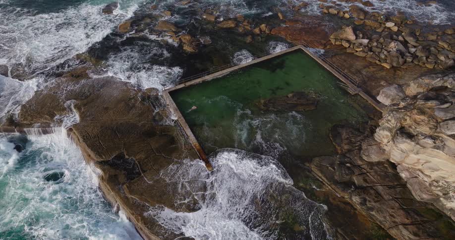 An aerial view of a natural rock pool filled with greenish water surrounded by rugged rocks and crashing ocean waves