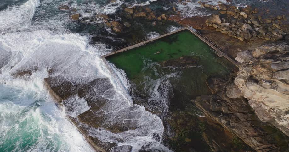 An aerial view of a natural rock pool filled with green water surrounded by rugged coastal rocks and waves crashing against the edges