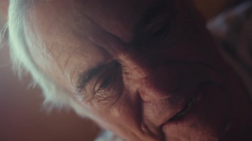 An elderly person with white hair is sitting indoors wearing a striped shirt with soft lighting illuminating the scene