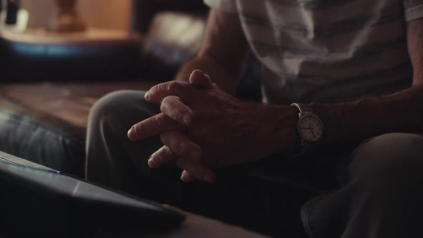 An elderly mans hands clasped together resting on his lap watching tablet