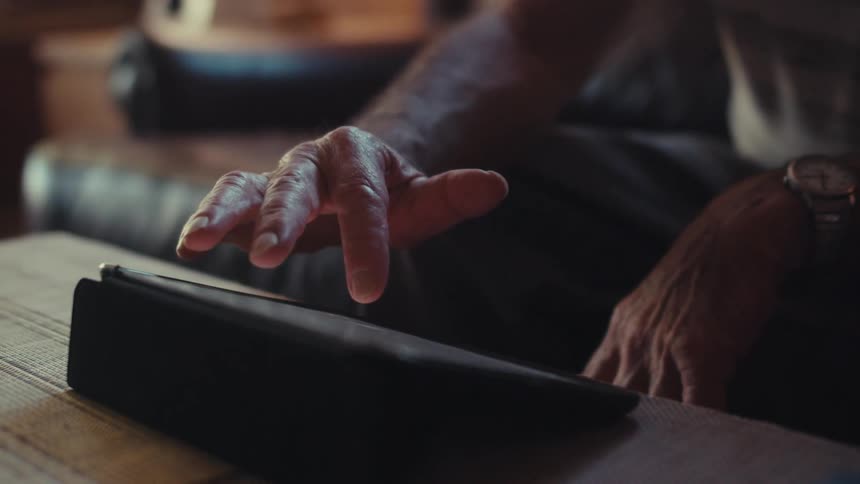 Elderly man sitting on the black couch uses a tablet in the living room