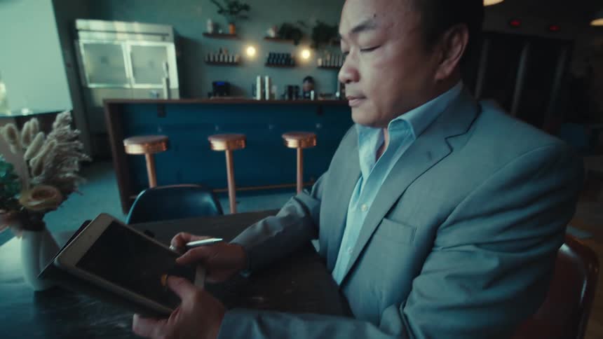 A person in a suit is sitting at a table in a modern cafe holding a tablet and a stylus