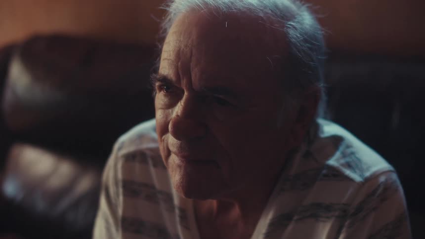 An elderly person with white hair is sitting indoors wearing a striped shirt with soft lighting illuminating the scene