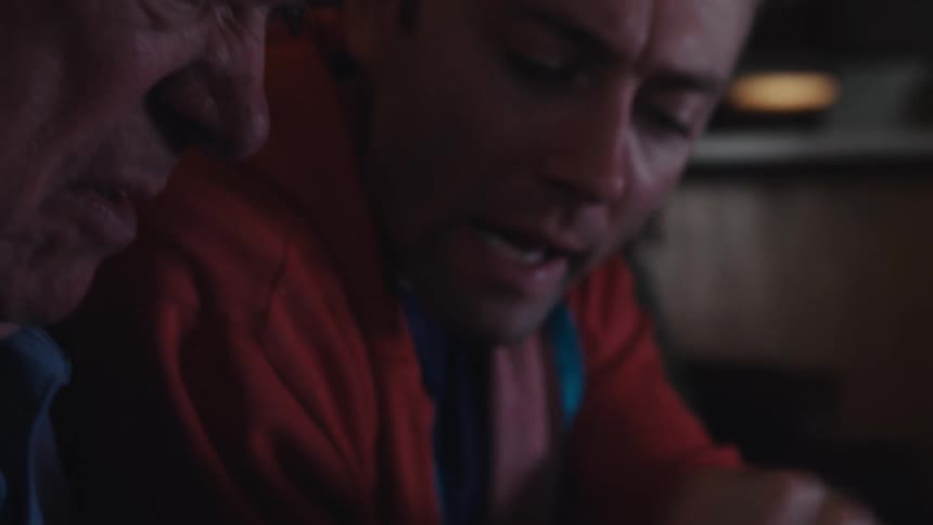 Father and son are sitting on a dark leather couch in a dimly lit living room playing video games
