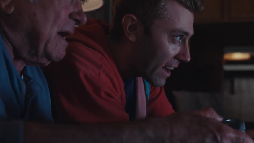 A young man and an elderly man sitting on a sofa and playing a video game looking intently ahead