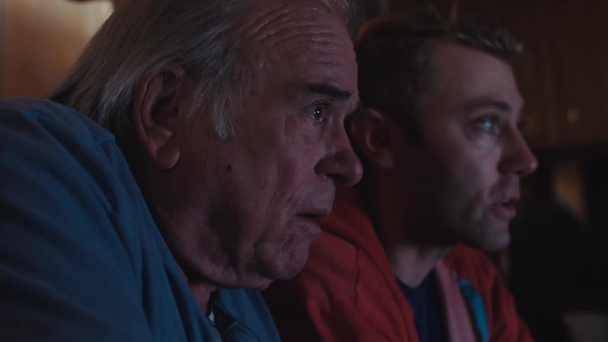 Father and son are sitting on a dark leather couch in a dimly lit living room playing video games